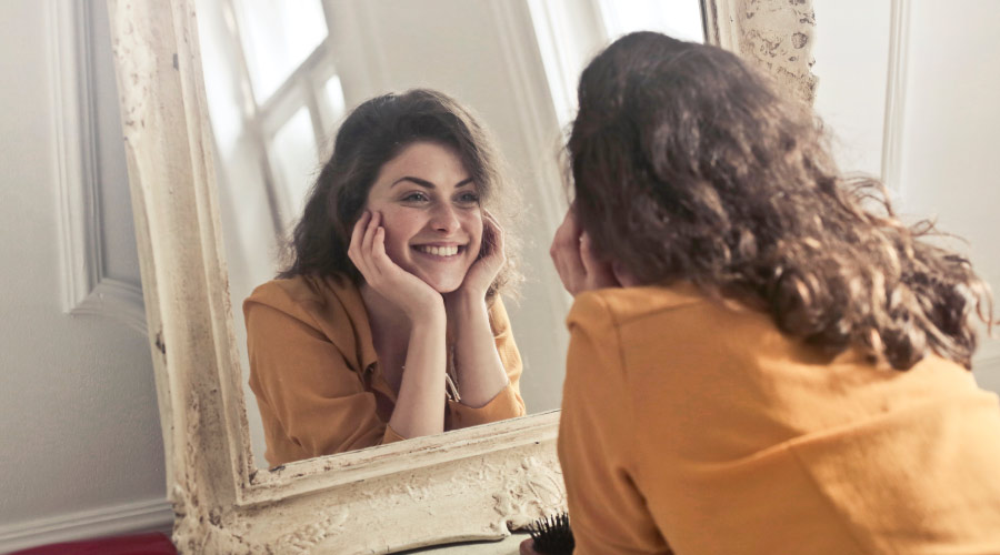 Brunette woman wearing a yellow blouse smiles in a mirror with an ornate border with her hands under her chin