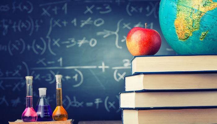 3 flasks of colored chemicals sit on a desk next to an apple and globe atop a stack of books against a chalkboard