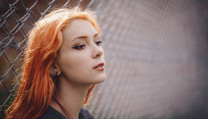Young woman with orange hair and a lip piercing sits against a chain-link fence
