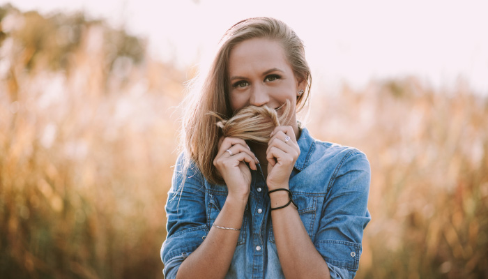 Blonde woman wearing a denim shirt covers her mouth with her hair due to bad breath while standing in a wheat field