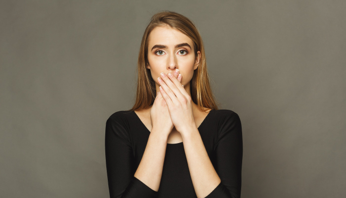 Blonde woman wearing a black shirt covers her mouth with her hands because she is embarrassed about her overbite