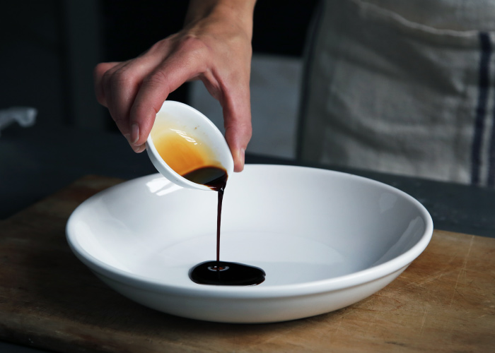 A hand pours out a small white container of balsamic vinegar into a shallow white bowl