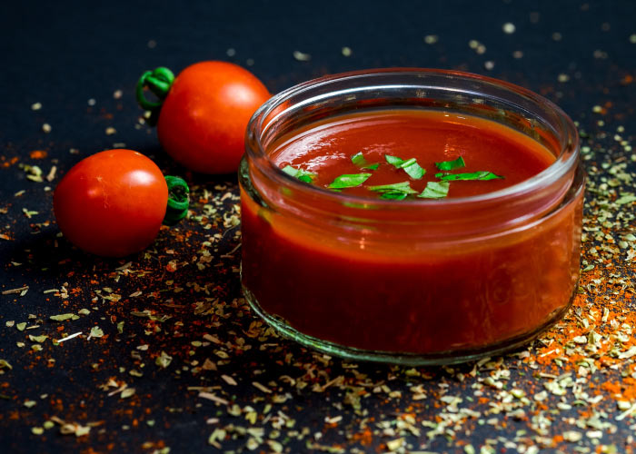 Clear container holds tomato sauce topped with basil next to 2 cherry tomatoes on a counter sprinkled with dried herbs